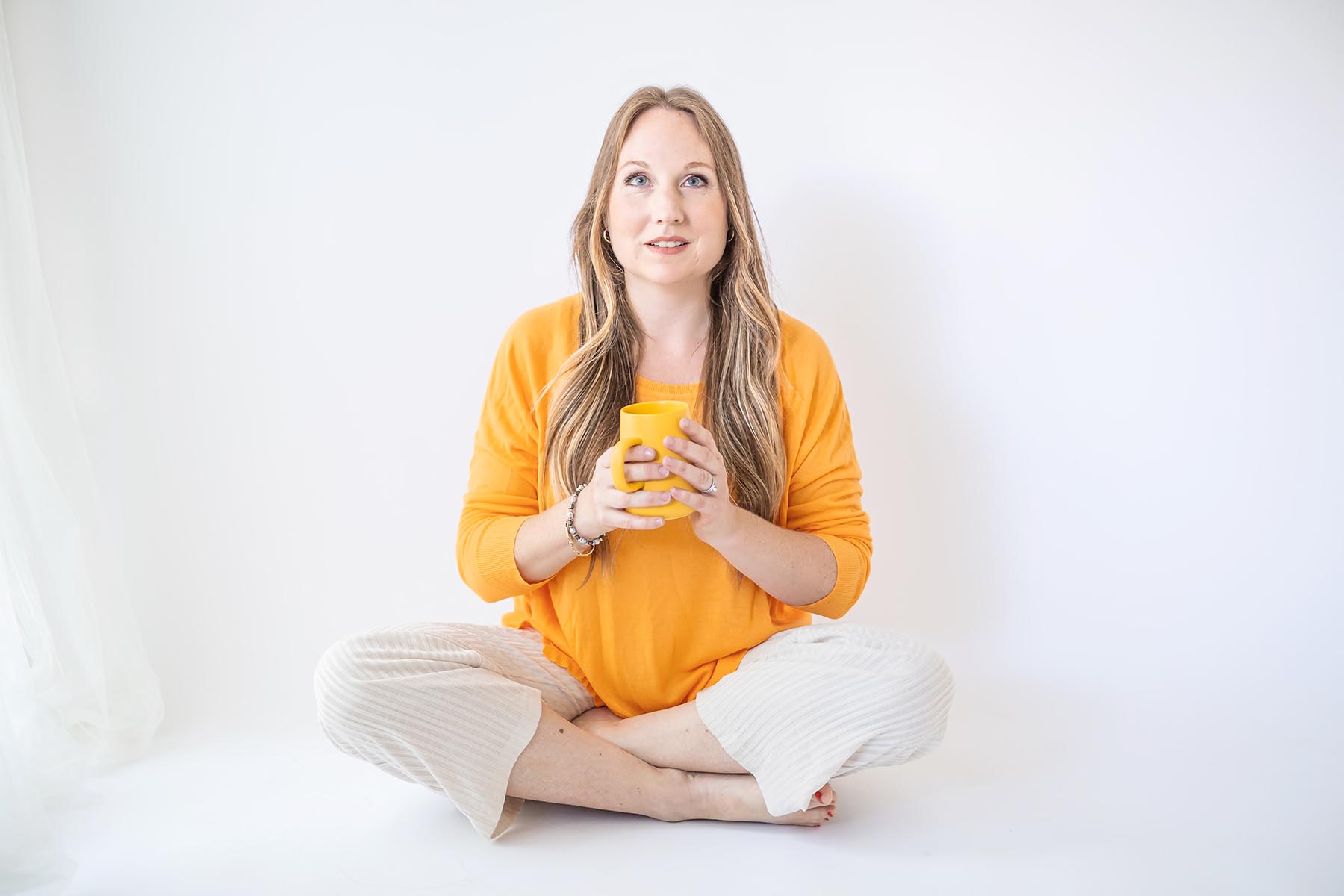 Founder of joybeforework.com, Rebecca Joy, cross-legged, mango-yellow long sleeve, holding yellow mug, looking up with hopeful smile